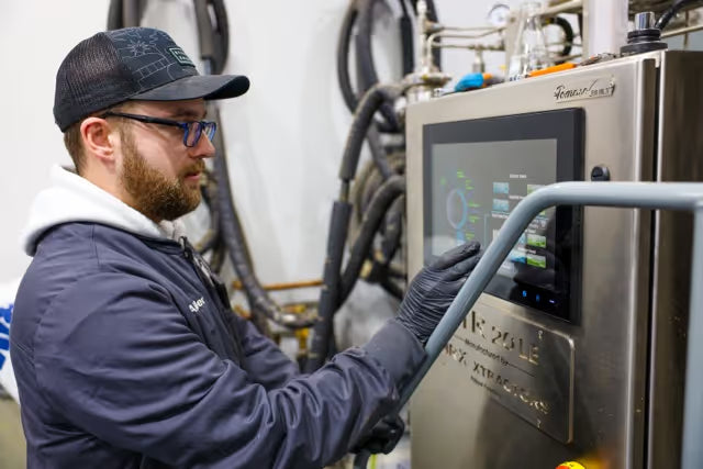 Man wearing glasses and a cap operating a touchscreen on industrial equipment in a factory setting.