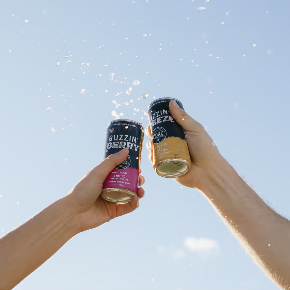 Two hands raising Buzzin Berry and Hydrate cans against a blue sky